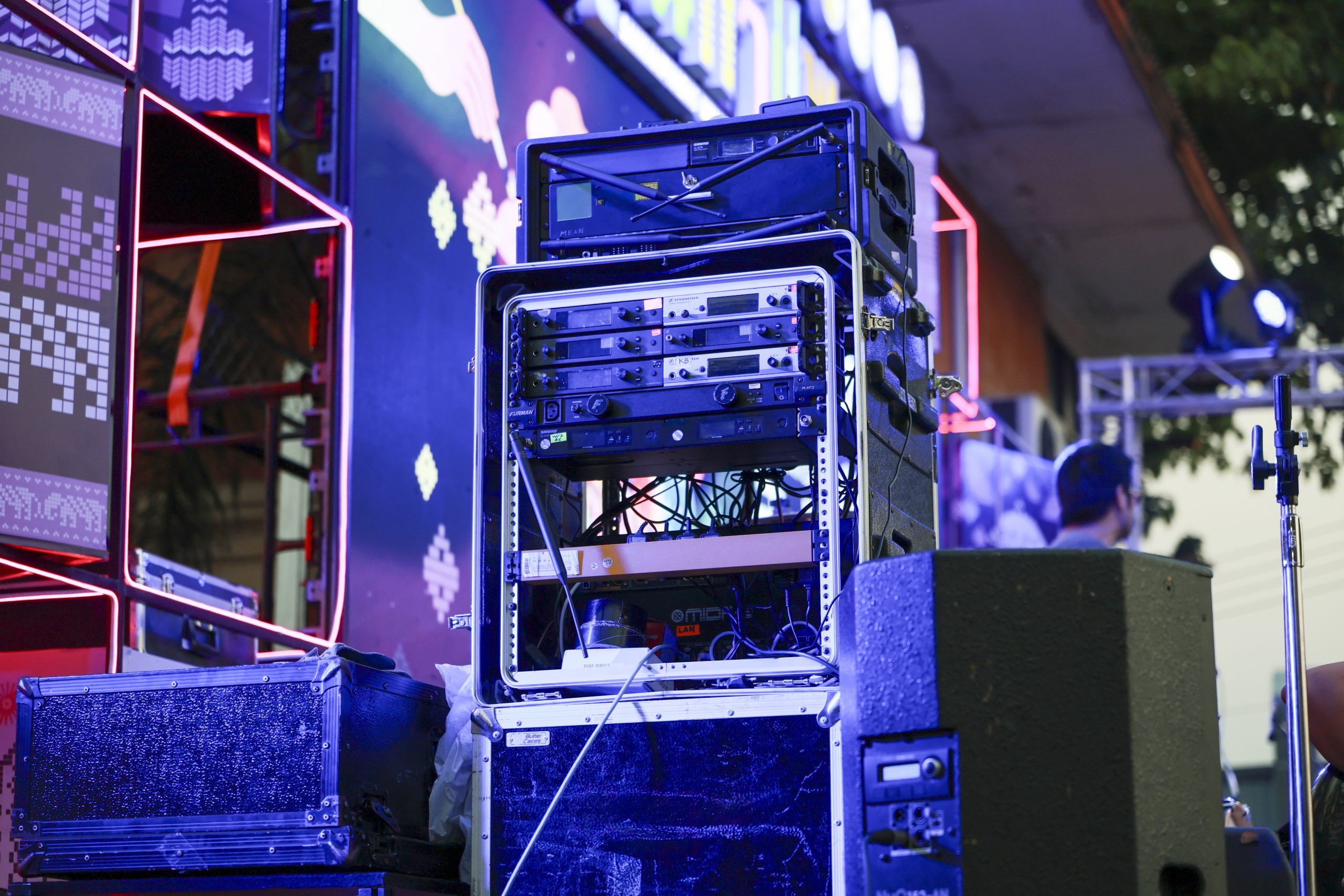 A rack-mounted audio equipment unit sits powered and connected with cables, feeding nearby speakers, on a lit stage with LED panels and colored neon lighting.