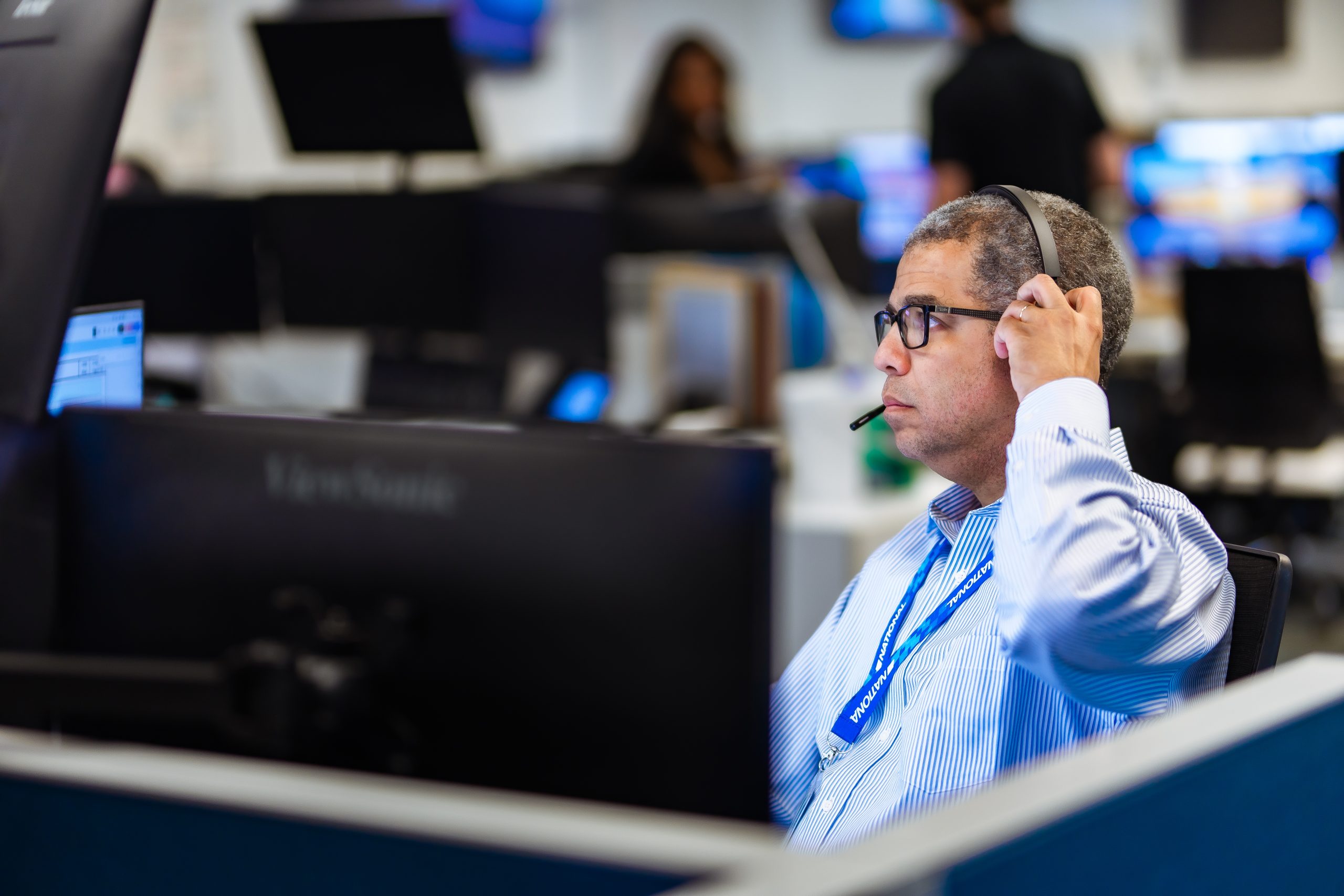 Man adjusts a headset while monitoring multiple computer screens, seated at a cubicle in a busy open-plan office with colleagues and blurred monitors in the background.