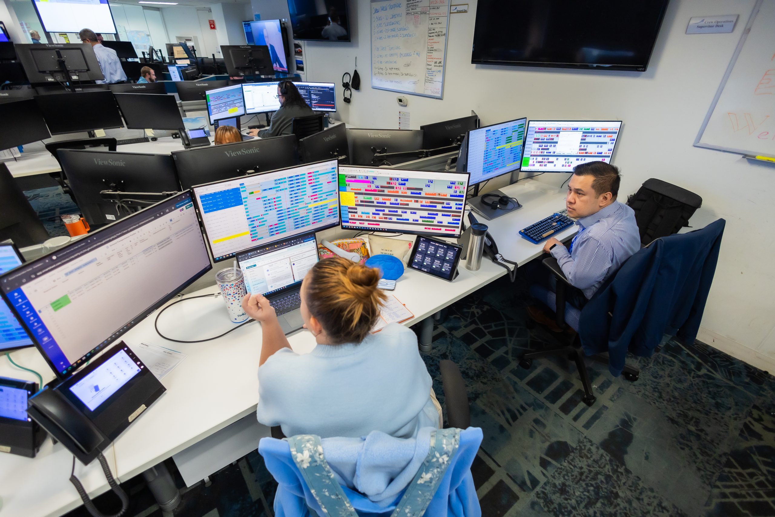 Multiple computer monitors display colorful schedules and data; two staff members type and discuss at a shared workstation in a brightly lit operations center.