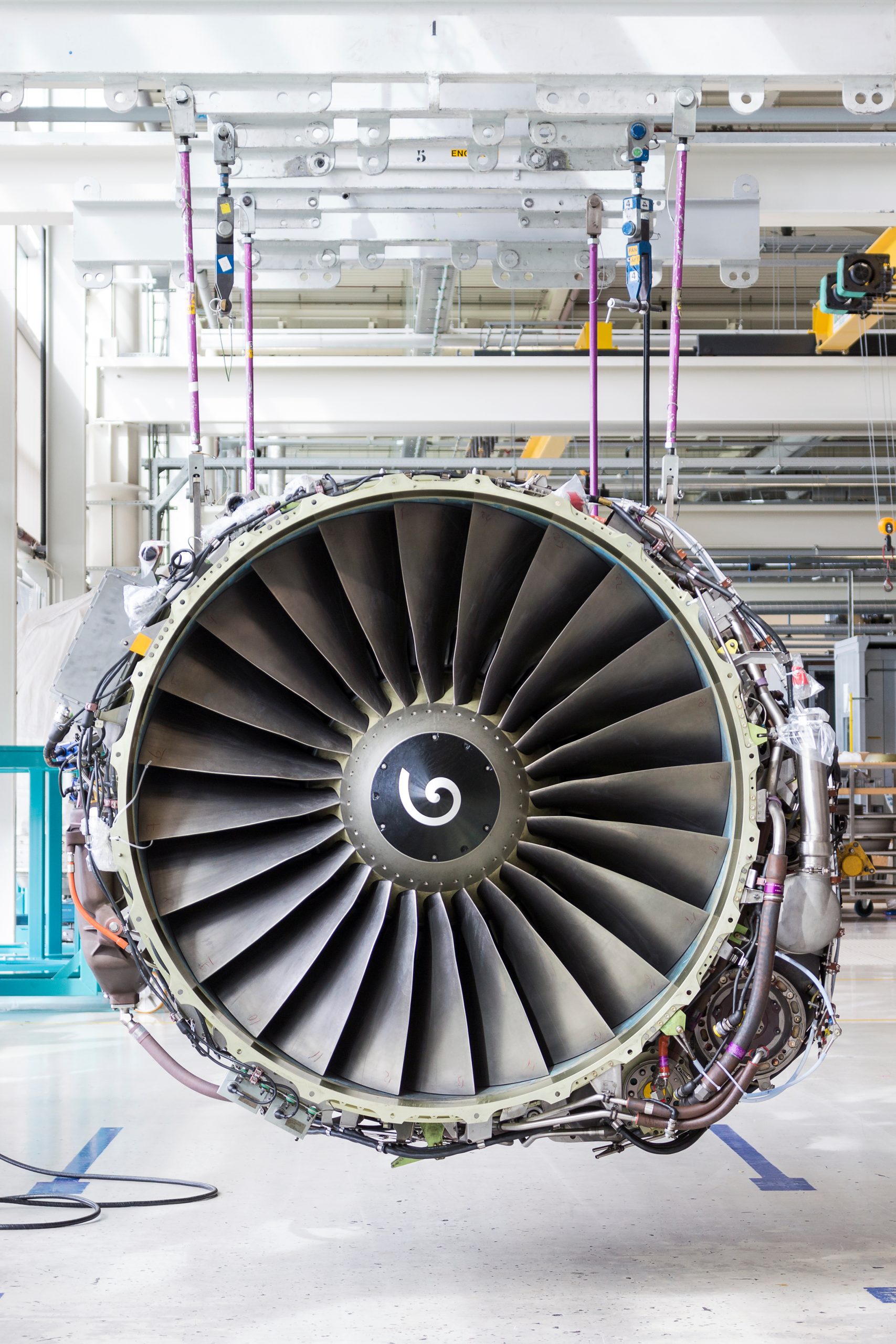 A large turbofan engine fan assembly hangs suspended from overhead rigging, its blades exposed and connected to cables and pipes inside a bright aircraft maintenance hangar.