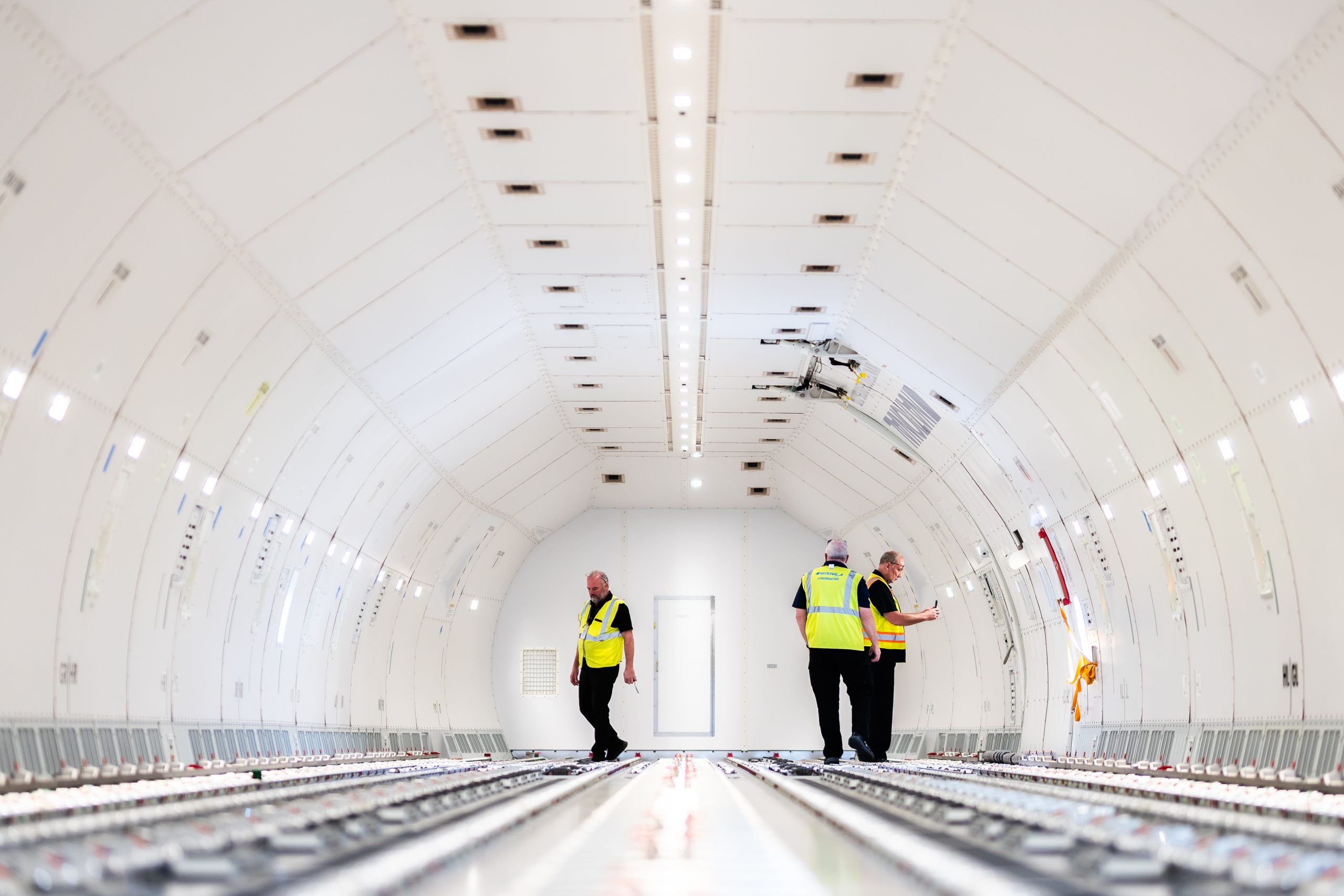 Three technicians walk and inspect an empty aircraft fuselage, standing on exposed floor tracks beneath a central row of lights and smooth white curved interior panels.
