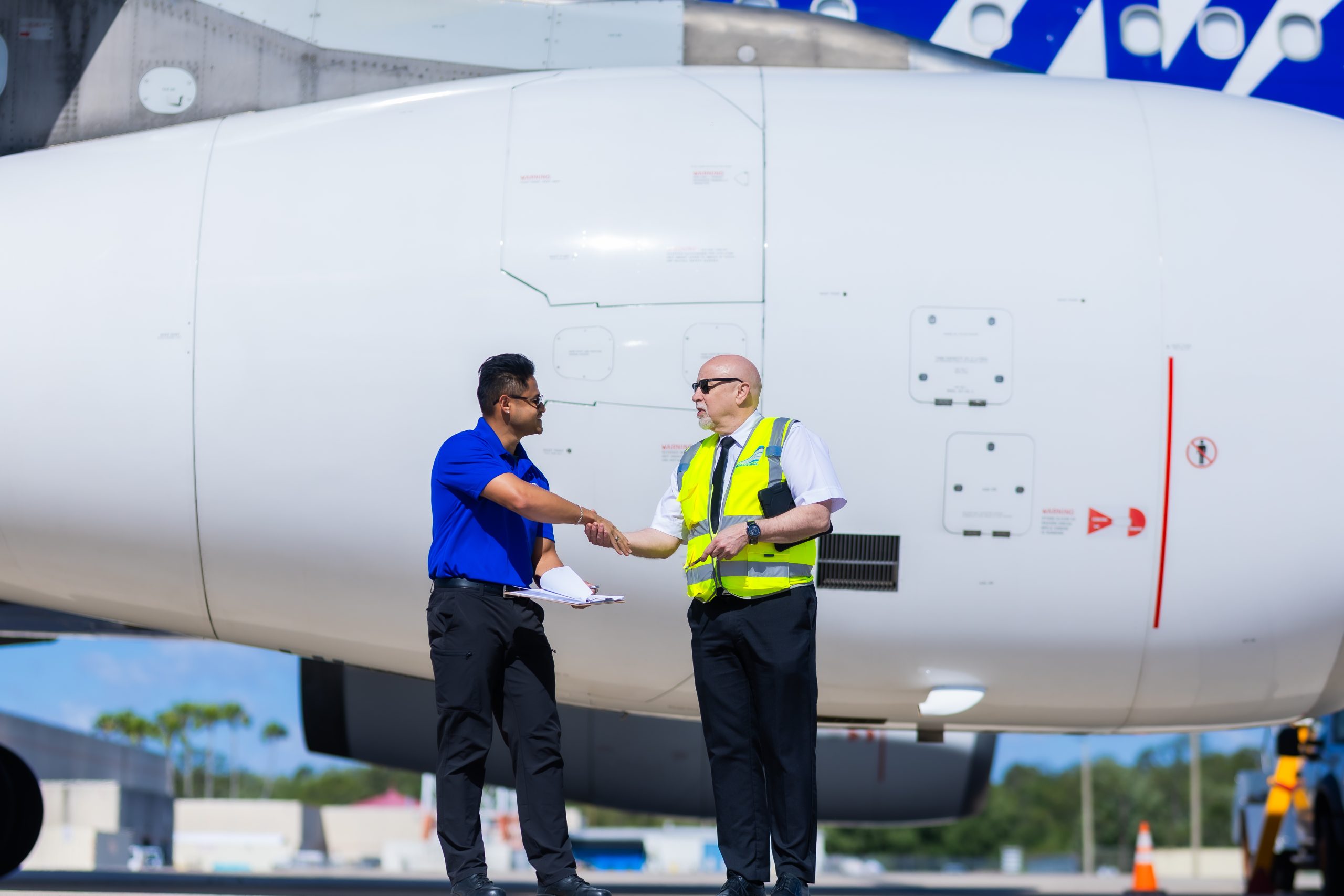 Two airport ground crew shake hands in front of a parked jet engine, one holding paperwork and the other wearing a reflective vest on a sunlit tarmac with safety cones nearby.