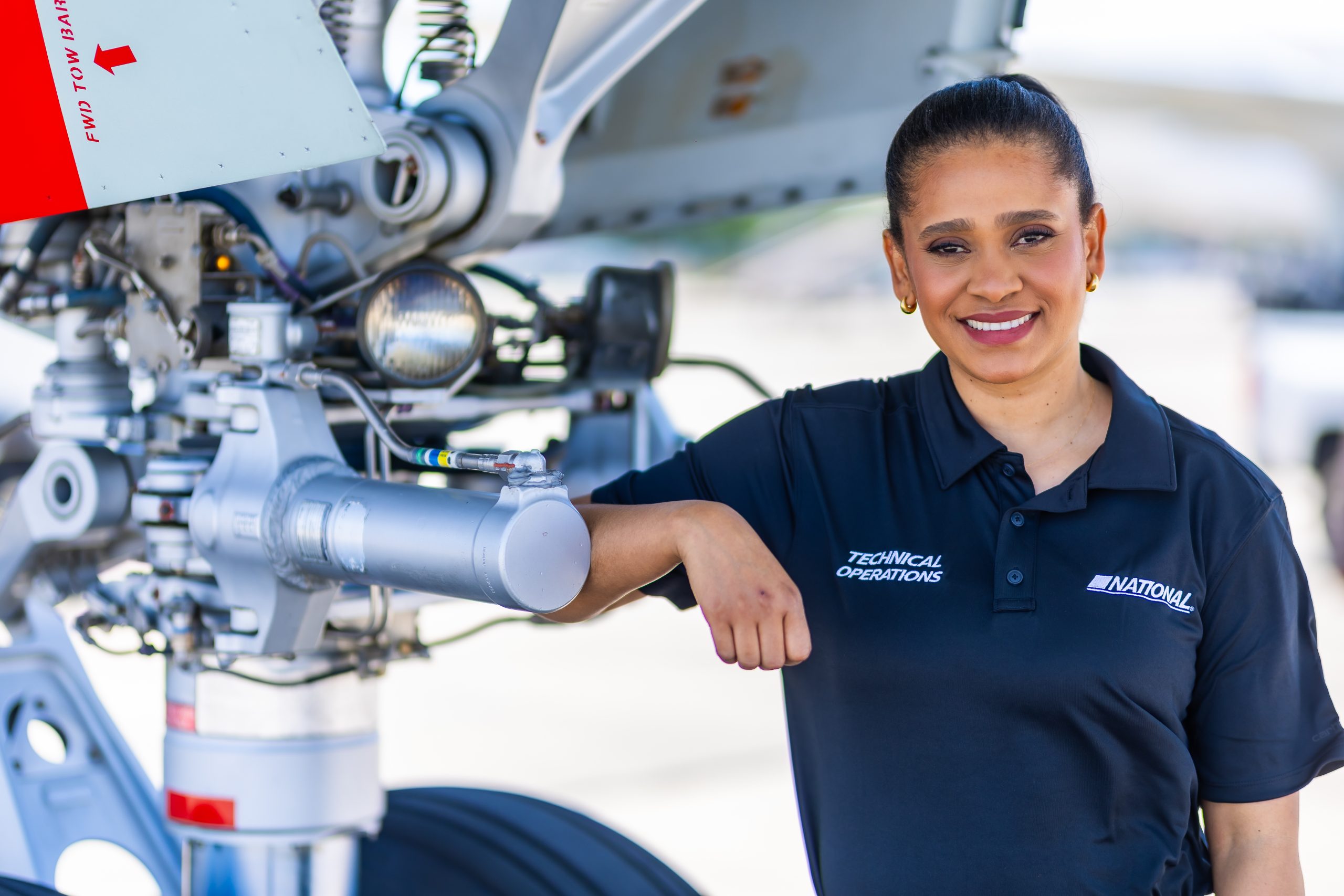 Woman technician leans on airplane landing gear, smiling while wearing a dark "Technical Operations" polo; aircraft mechanical components and a blurred tarmac form the background.
