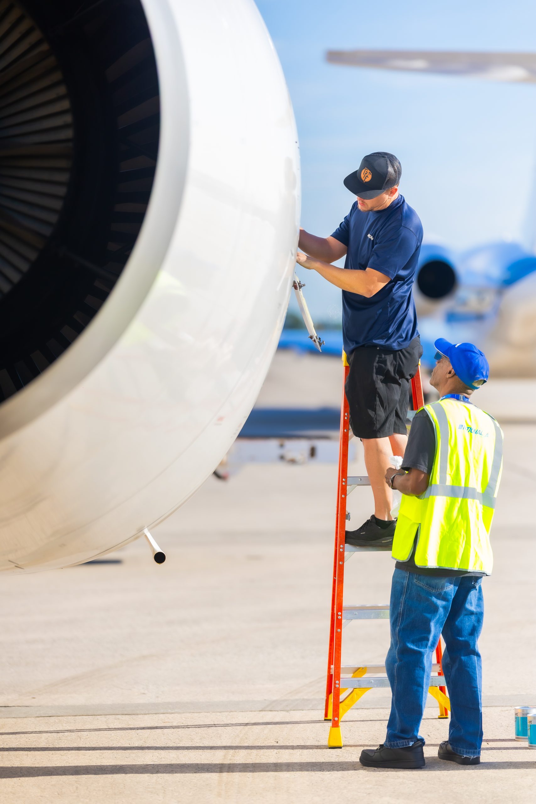 Jet engine — technician on a red ladder inspects and adjusts an exterior panel while a colleague in a high-visibility vest watches on a sunny airport tarmac with other aircraft nearby.