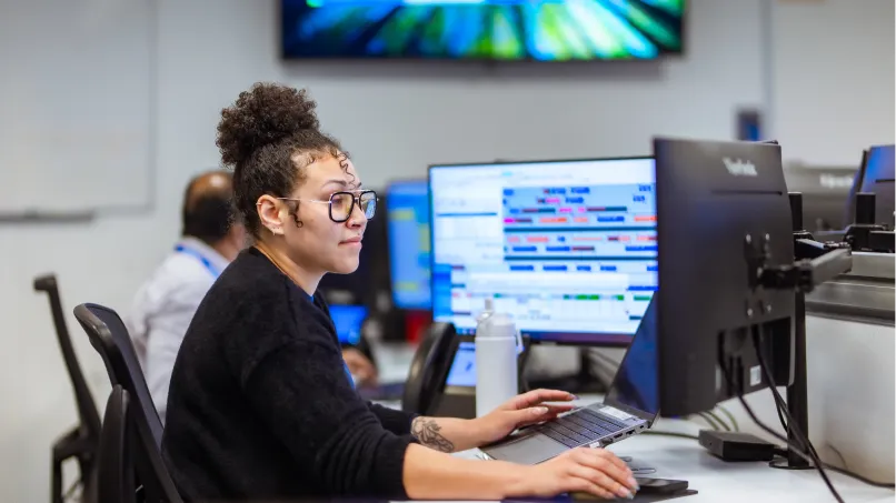 A person at a desk typing on a laptop while glancing at multiple large monitors; several screens display colorful schedules, and colleagues and office equipment fill the open-plan workspace.