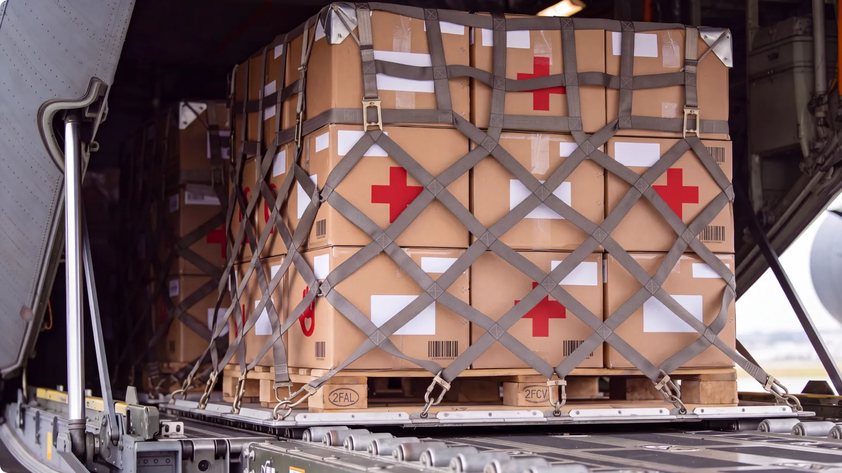 Stacks of cardboard medical supply crates, marked with red crosses, are strapped and palletized under cargo nets inside an aircraft hold for air transport.