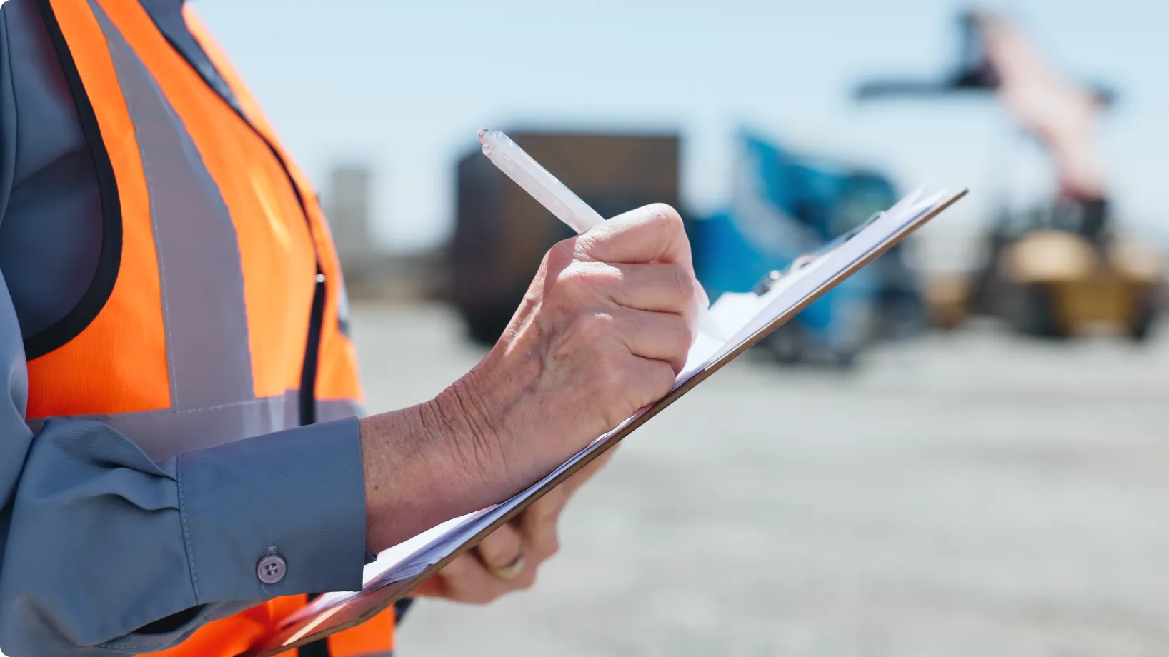 Clipboard being written on by a worker wearing an orange safety vest at a dusty construction site, with blurred heavy machinery and shipping containers visible in the background.