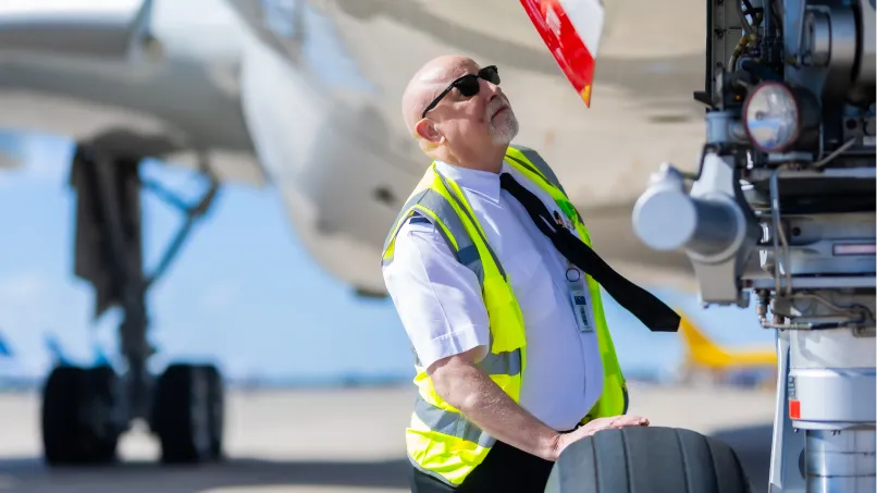 Ground crew member in a high-visibility vest inspects an aircraft's landing gear and tire, looking upward on a sunlit airport tarmac with other planes blurred behind.