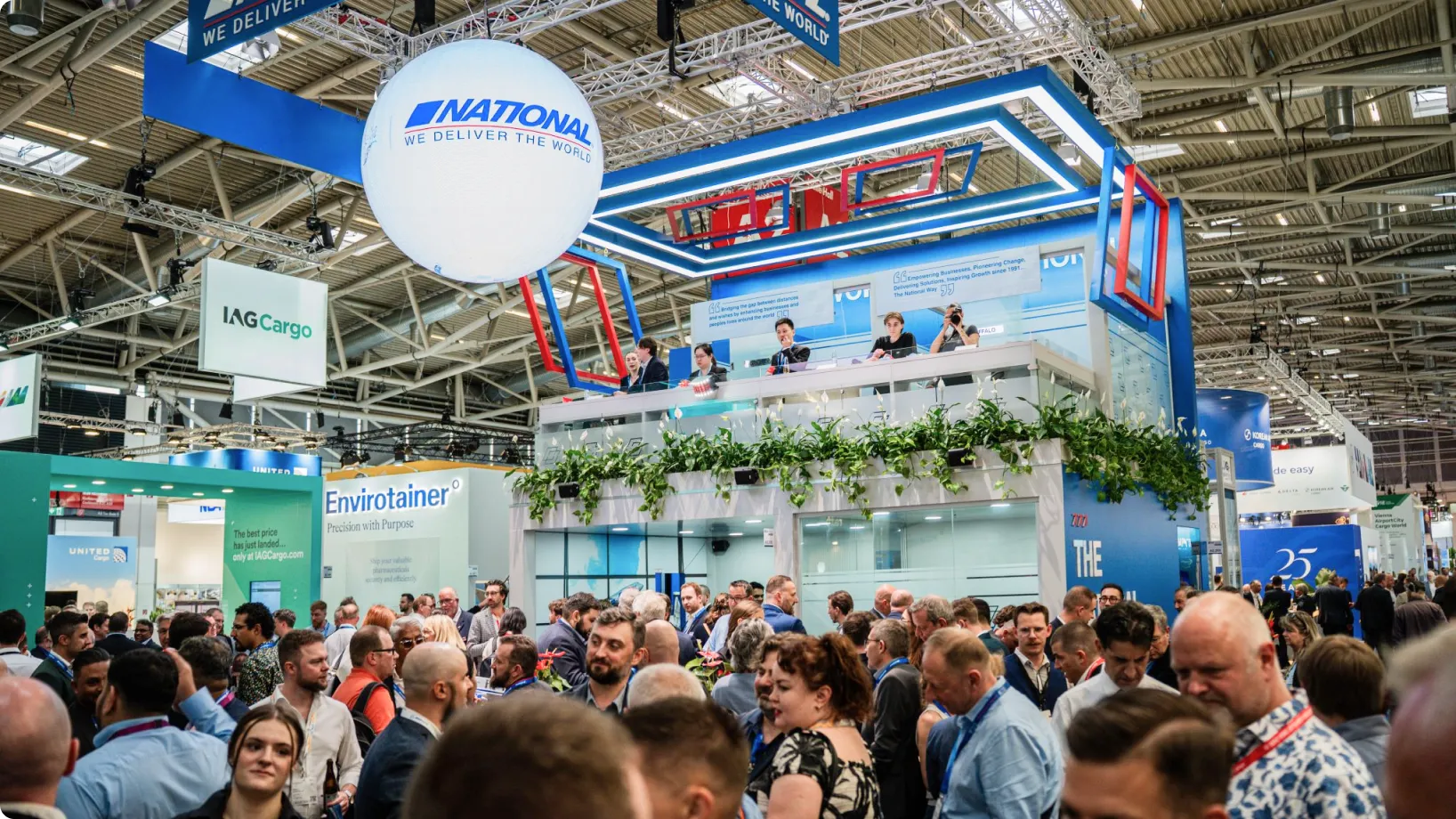 Large double-decker exhibition booth (branded "National") hosts staff on an upper platform while a dense crowd mills and converses below amid a busy, well-lit convention hall with overhead signage.