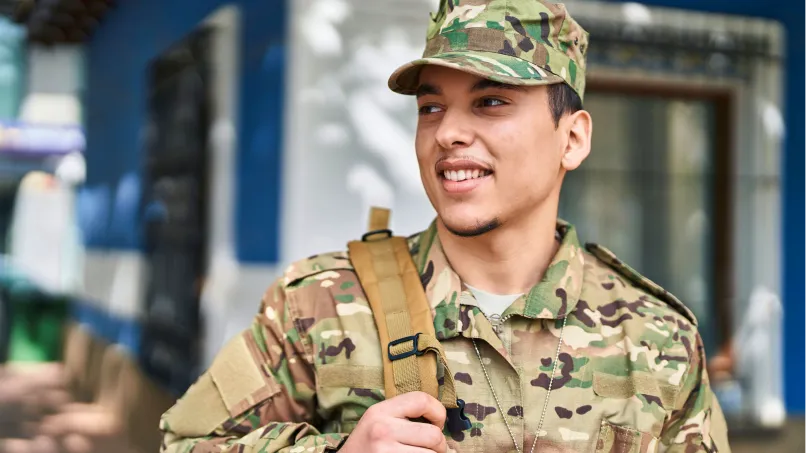 A smiling soldier adjusts a backpack strap while standing outside a blue-and-white building, wearing camouflage uniform and cap, and looking off-camera to his left.