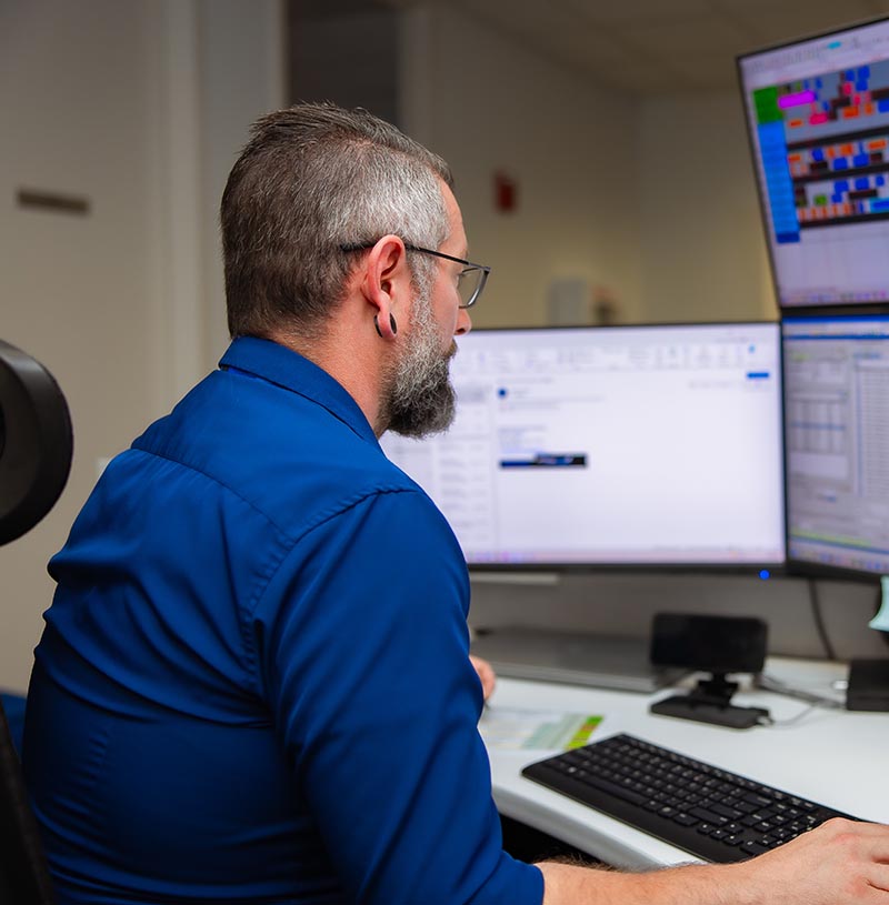 A man with glasses and a gray beard sits at a curved desk, typing and viewing multiple monitors showing spreadsheets and charts in a brightly lit office workspace.