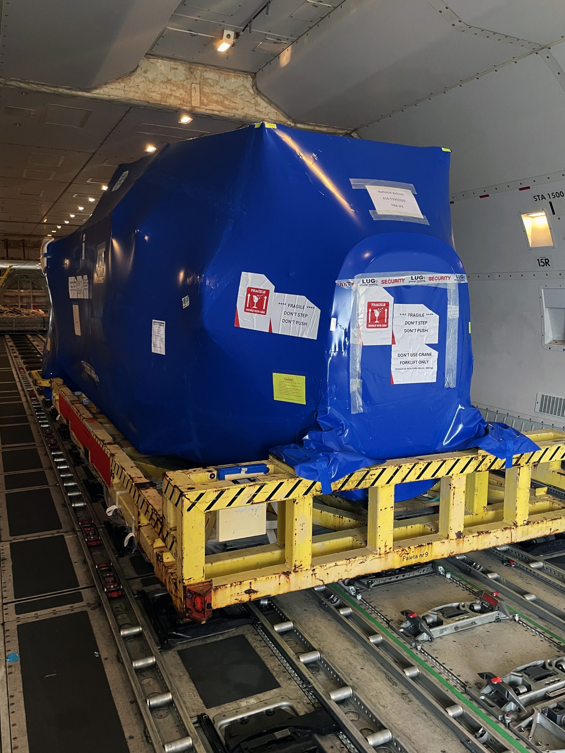 Large blue-wrapped cargo unit sits on a yellow metal pallet, labeled fragile and secured, inside an aircraft cargo hold atop roller tracks beneath overhead lighting.
