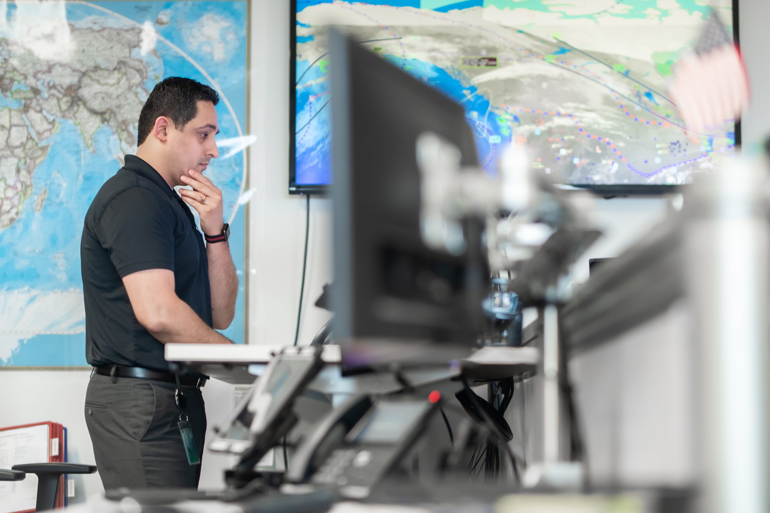 Person at a standing desk studies a computer monitor, hand on chin in thought, surrounded by telephones and large world-map and weather displays on the wall, in an office setting.
