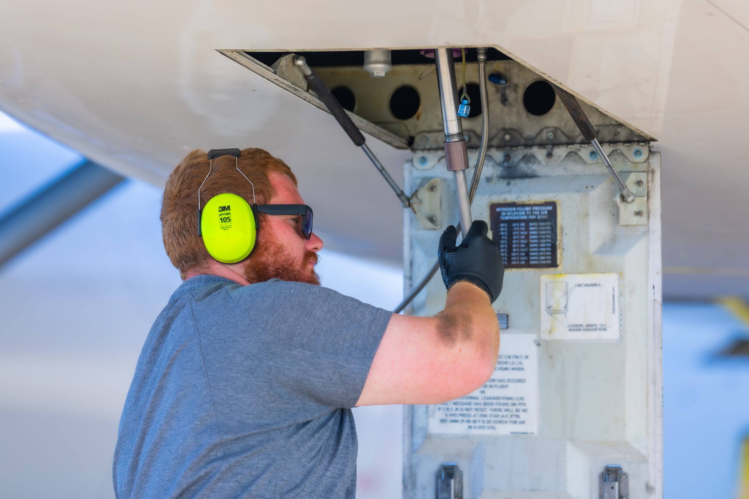 Technician wearing neon ear protection, gloves and sunglasses tightens or inspects wiring inside an open aircraft access panel beneath the fuselage on a sunny tarmac, using a handheld tool.