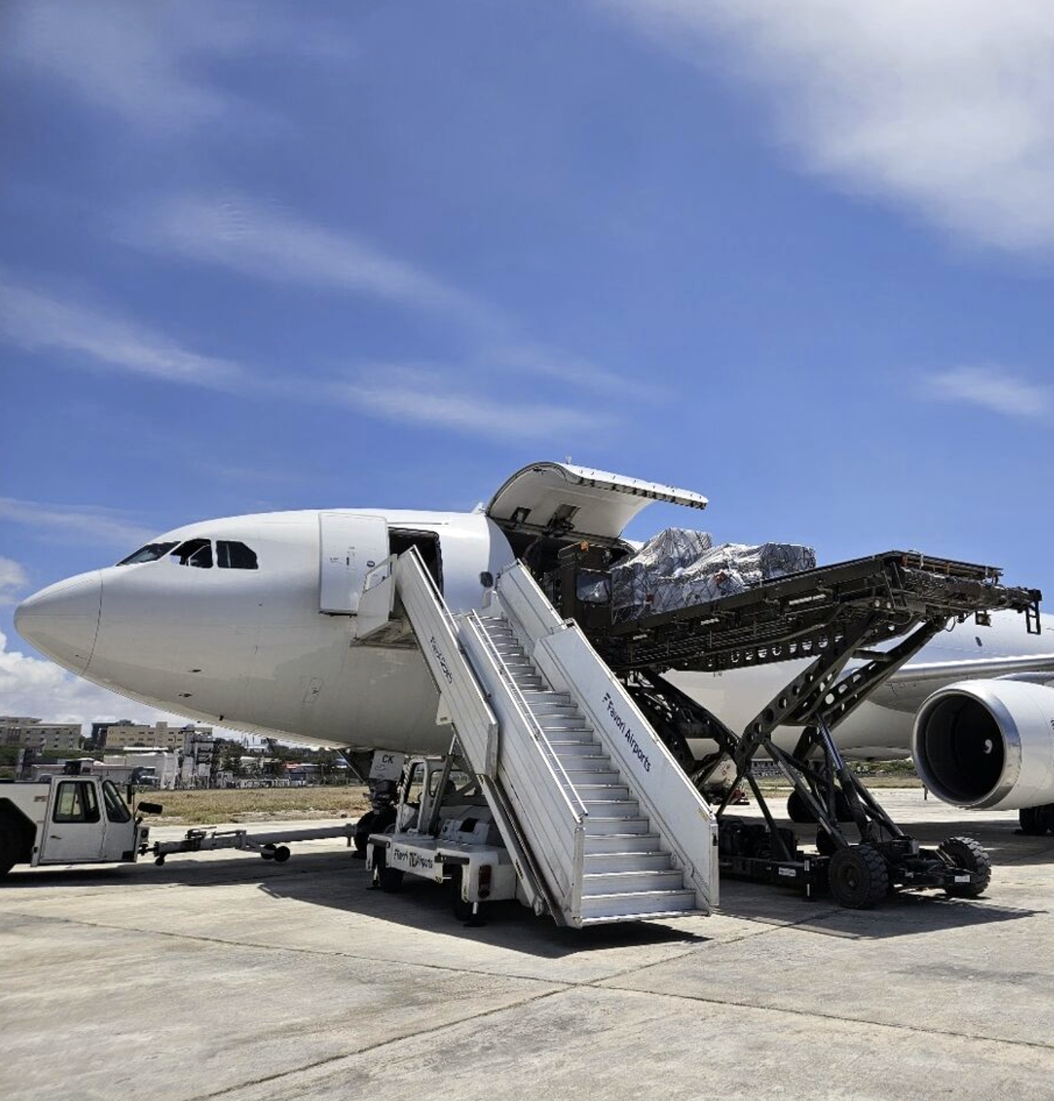 Wide-body airliner; forward cargo door open as a high-loader lifts palletized freight into the hold while mobile stairs and ground vehicles stand on a sunlit airport apron under a blue sky.